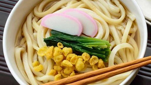 Bowl of udon noodles with fish cake slices, green vegetables, corn, and wooden chopsticks.