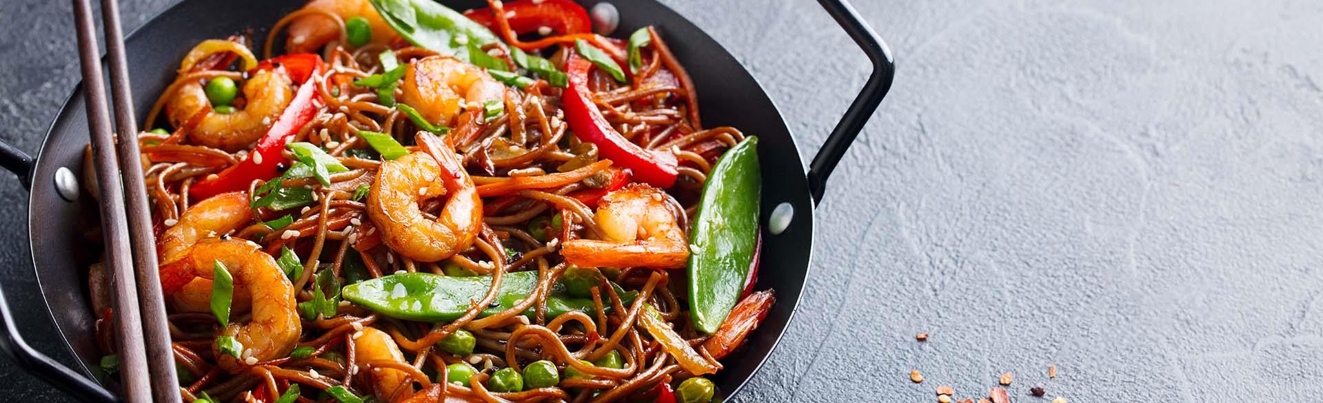Stir-fried noodles with shrimp, vegetables, and herbs in a black wok, with chopsticks and sesame seeds nearby.