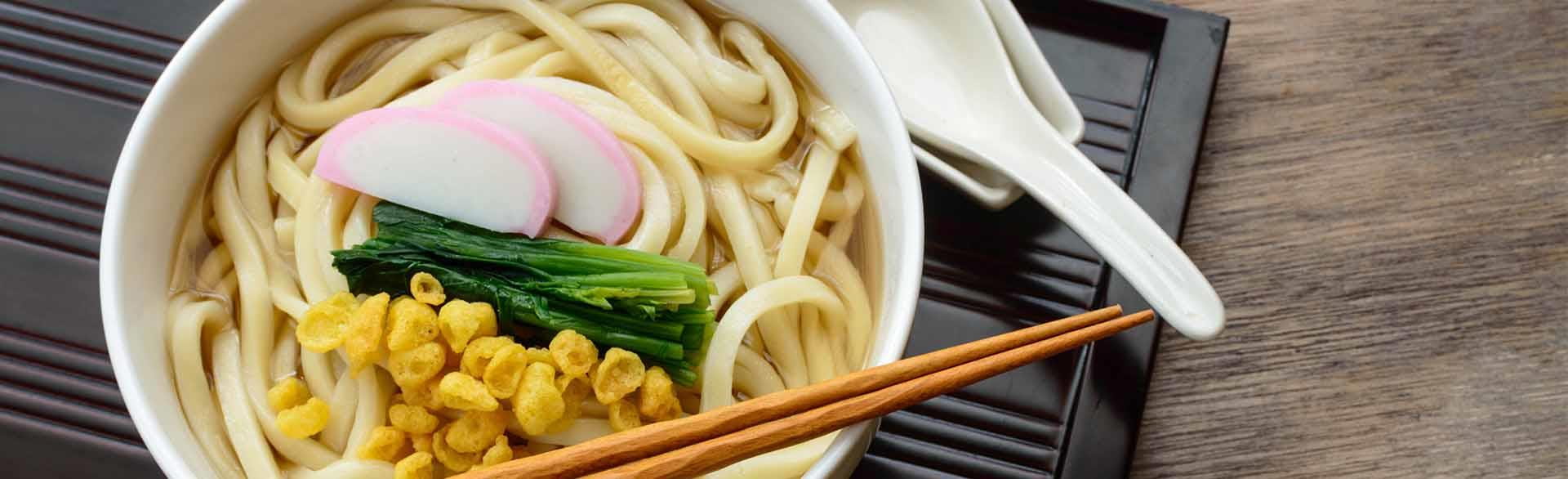 Bowl of noodle soup with fish cake slices, leafy greens, corn, chopsticks, and a ceramic spoon.