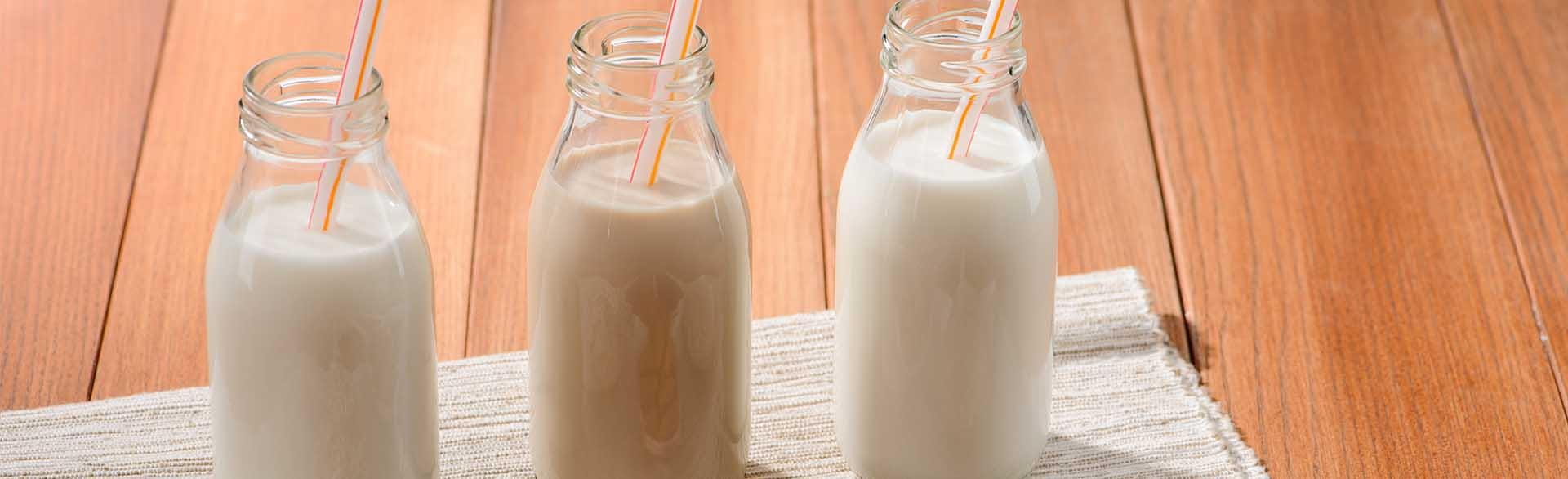 Three glass bottles of milk with straws, including chocolate and white milk, on a wooden surface.