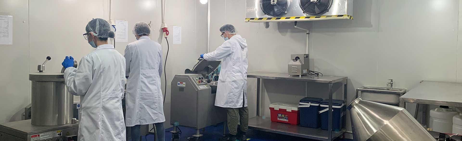 Three lab workers in protective gear operating equipment and standing near large stainless steel containers.